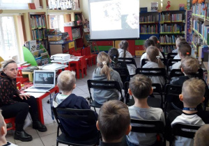 Grupa uczniów siedzi w sali bibliotecznej i ogląda prezentację multimedialną