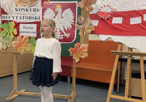 Uczennica w galowym stroju na tle ozdobnej tablicy z napisem Konkurs recytatorski - wiersze patriotyczne