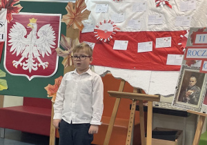 Uczeń w galowym stroju na tle ozdobnej tablicy z napisem Konkurs recytatorski - wiersze patriotyczne
