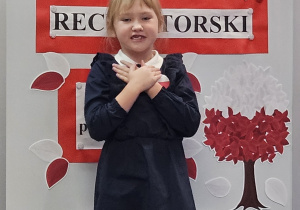 Uczennica w galowym stroju na tle ozdobnej tablicy z napisem Konkurs recytatorski - wiersze patriotyczne.