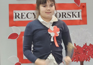Uczennica w galowym stroju na tle ozdobnej tablicy z napisem Konkurs recytatorski - wiersze patriotyczne.