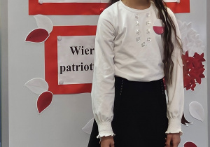 Uczennica w galowym stroju na tle ozdobnej tablicy z napisem Konkurs recytatorski - wiersze patriotyczne.