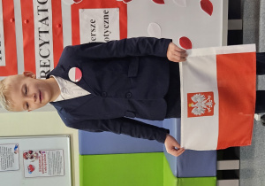 Uczeń w galowym stroju na tle ozdobnej tablicy z napisem Konkurs recytatorski - wiersze patriotyczne.