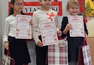 Trzy uczennice w strojach galowych z dyplomami i torbami upominkowymi w dłoniach na tle ozdobnej tablicy z napisem Konkurs recytatorski - wiersze patriotyczne.
