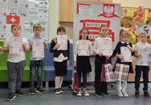 Grupa dzieci w galowych strojach z dyplomami w dłoniach na tle ozdobnej tablicy z napisem Konkurs recytatorski - wiersze patriotyczne.