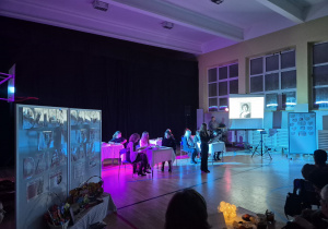 Występ uczennicy, w tle nauczyciele i uczniowie siedzący przy stolikach, na ekranie prezentacja o polskich poetach