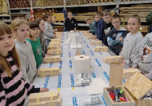 Uczniowie stoją przy stołach, na których leżą elementy desek.