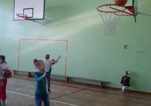 Dzieci rzucają piłkę do kosza w sali gimnastycznej.