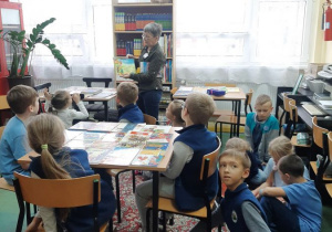 Grupa dzieci, siedzących przy stoliku i na dywanie, słucha opowiadania nauczyciela.