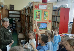 Dzieci stoją przed nauczycielem prowadzącym zajęcia w bibliotece szkolnej.