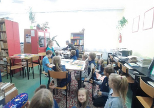 Uczniowie siedzą na dywanie i krzesłach w bibliotece szkolnej.