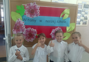 Uśmiechnięte pierwszoklasistki pozują do zdjęcia przy tablicy z napisem "Witamy w naszej szkole".