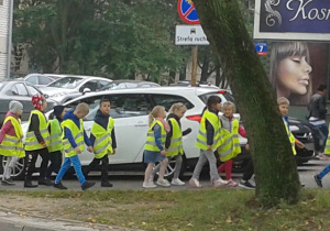Grupa uczniów w kamizelkach odblaskowych, idąca parami w kolumnie, po chodniku.