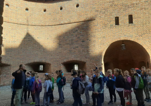 Dzieci słuchają opowieści przewodnika o historii związanej z murami Starego Miasta.