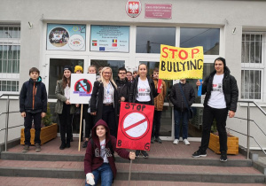 Grupa uczniów trzymających transparenty stoi wraz z koordynatorem projektu przed budynkiem szkoły.