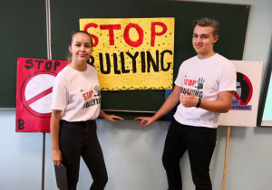 Dwoje uśmiechniętych uczniów w koszulkach z napisem: STOP BULLYING stoją na tle tablicy z plakatem.