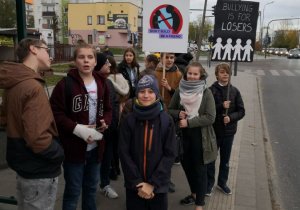 Grupa uczniów trzymających transparenty stoi na przystanku autobusowym.