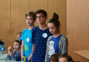 Troje uczniów, dwóch chłopców i dziewczynka, stoją przy stole dyskusyjnym przedstawiając wybrane prawa dziecka.