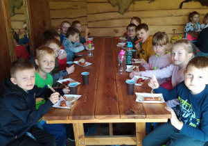 Dzieci siedzą przy stołach i jedzą kiełbaski z grilla
