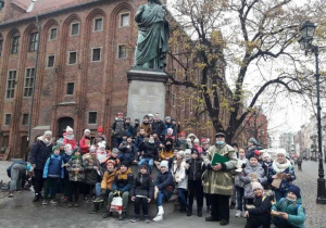 Grupa dzieci wraz z panem przewodnikiem ubranym w zielony płaszcz, pozuje do zdjęcia pod pomnikiem Mikołaja Kopernika