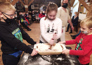 Dwóch chłopców trzyma gliniane naczynie, a dziewczynka miesza składniki potrzebne do ciasta na pierniki
