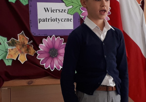 Uczeń w galowym stroju na tle ozdobnej tablicy z napisem Konkurs recytatorski - wiersze patriotyczne.