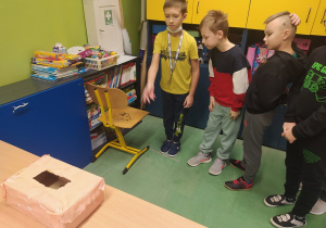 Czterej chłopcy rzucają monetami do papierowego pudełka, które stoi na ławce przed nimi.