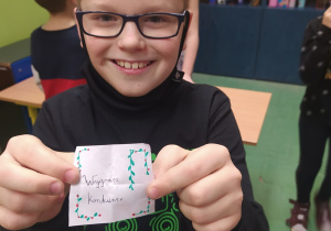 Uśmiechnięty chłopiec w okularach trzyma w ręku kartkę ze swoją wróżbą "Wygrasz konkurs"