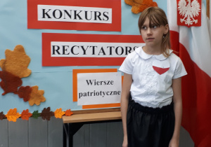 Uczennica w galowym stroju na tle ozdobnej tablicy z napisem Konkurs recytatorski - wiersze patriotyczne