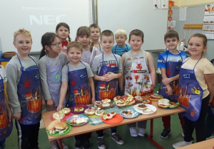Grupa uczniów w fartuszkach prezentuje przygotowane przez siebie kanapki.