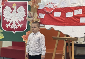 Uczeń w galowym stroju na tle ozdobnej tablicy z napisem Konkurs recytatorski - wiersze patriotyczne